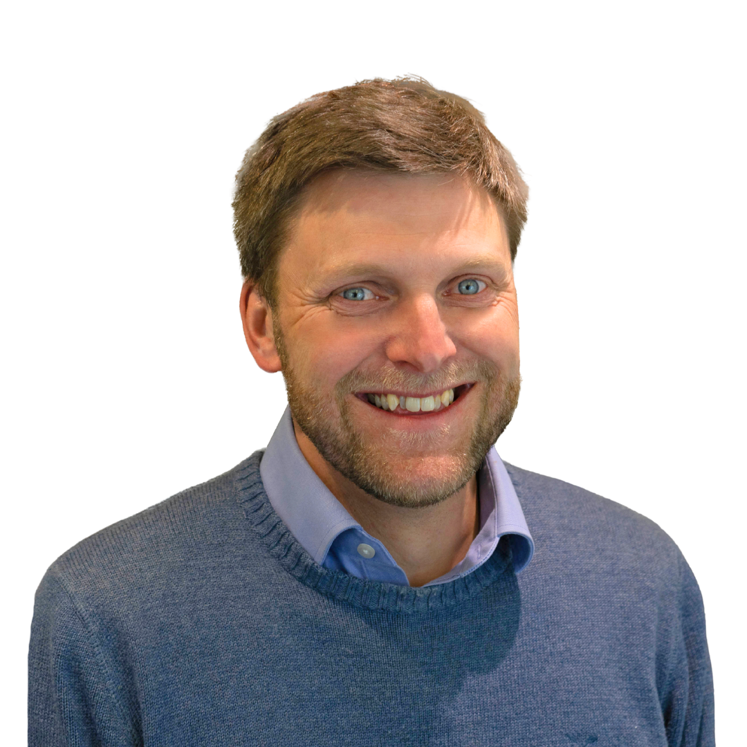 A man with short brown hair and a beard, wearing a blue sweater over a collared shirt, smiling at the camera against a plain background.