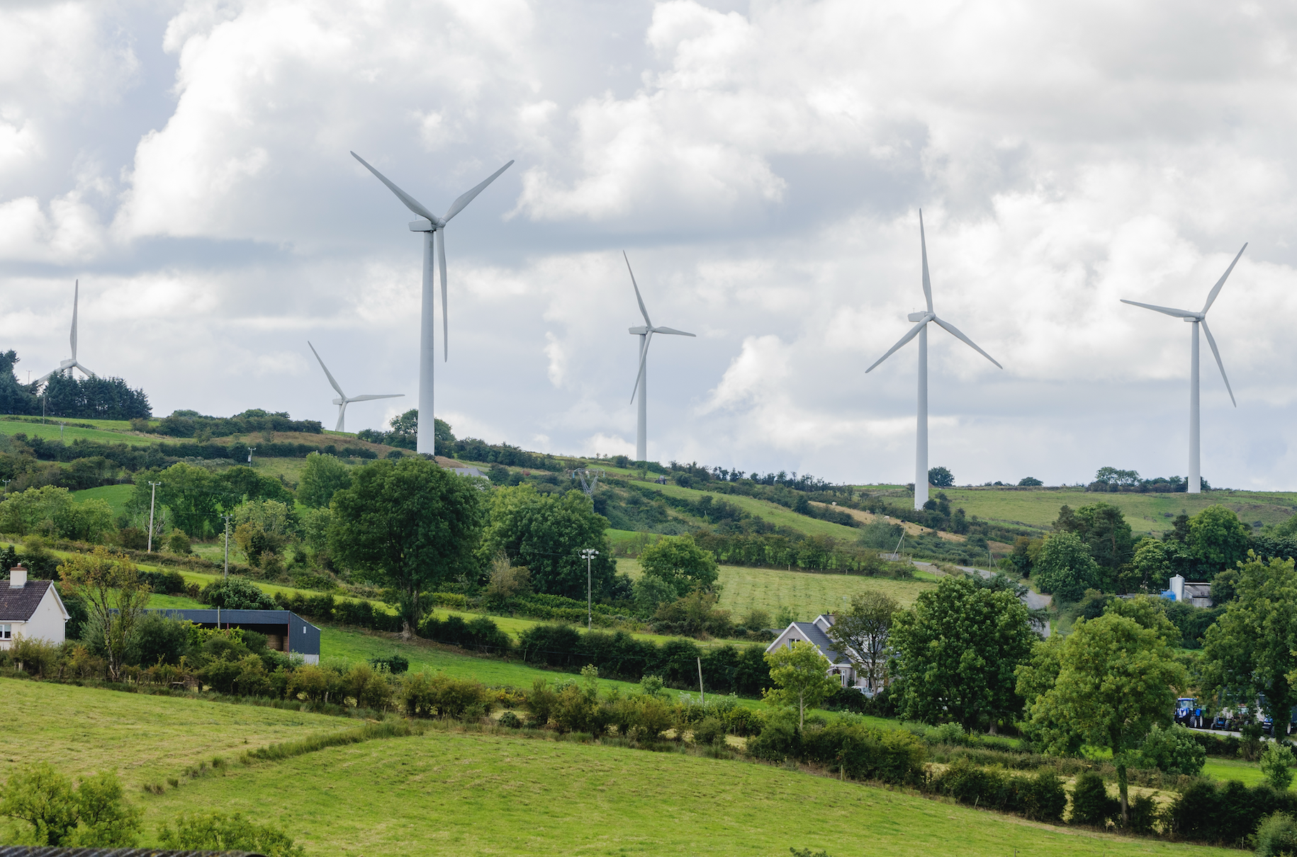 A landscape view featuring several wind turbines on rolling green hills, with trees and houses in the foreground under a partly cloudy sky.