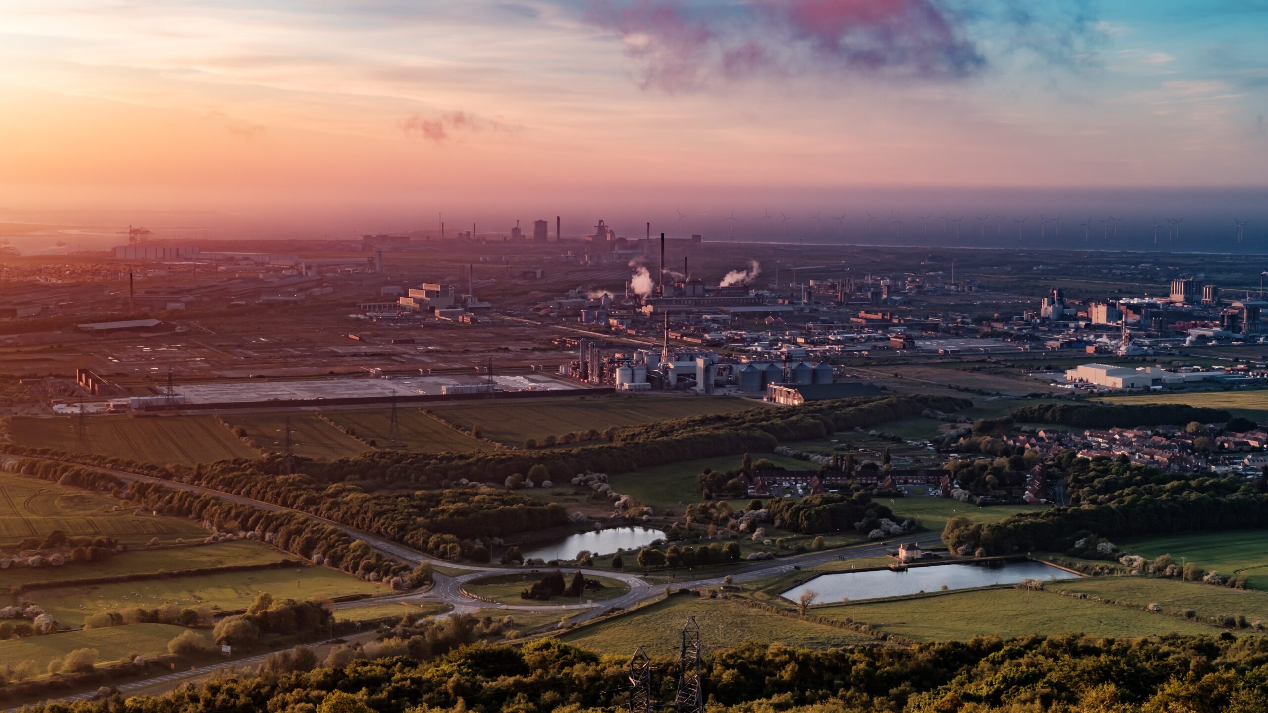 A panoramic view of an industrial landscape during sunset, featuring factories and smokestacks in the background, surrounded by green fields and bodies of water.