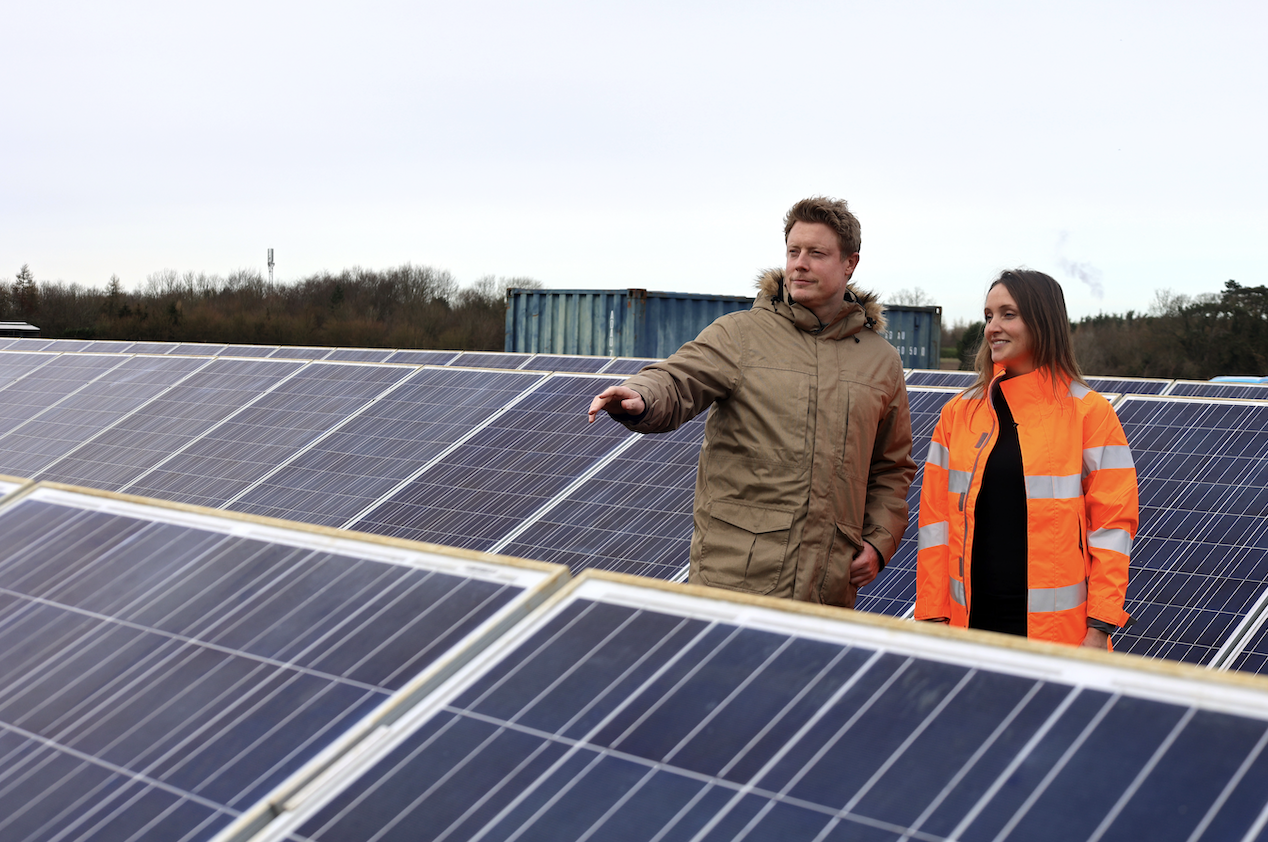 A man and a woman discussing solar panels on a photovoltaic installation. The man, wearing a beige jacket, points at the panels, while the woman, dressed in an orange safety jacket, observes. A cloudy sky is visible in the background.