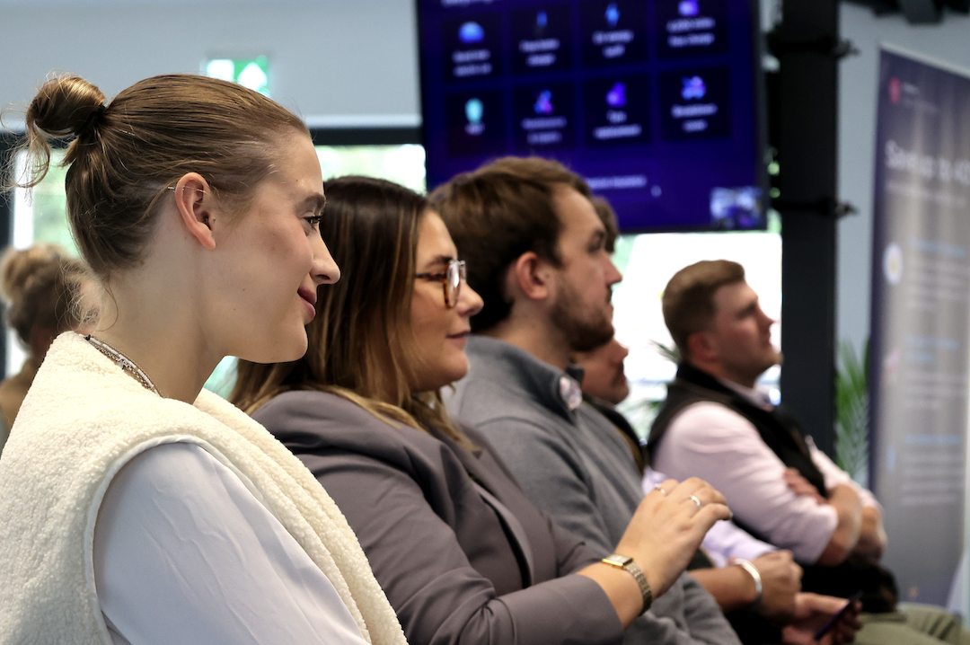 A group of professionals attentively listening during a presentation in a modern office setting. The focus is on three individuals in the foreground, with a screen displaying information in the background.