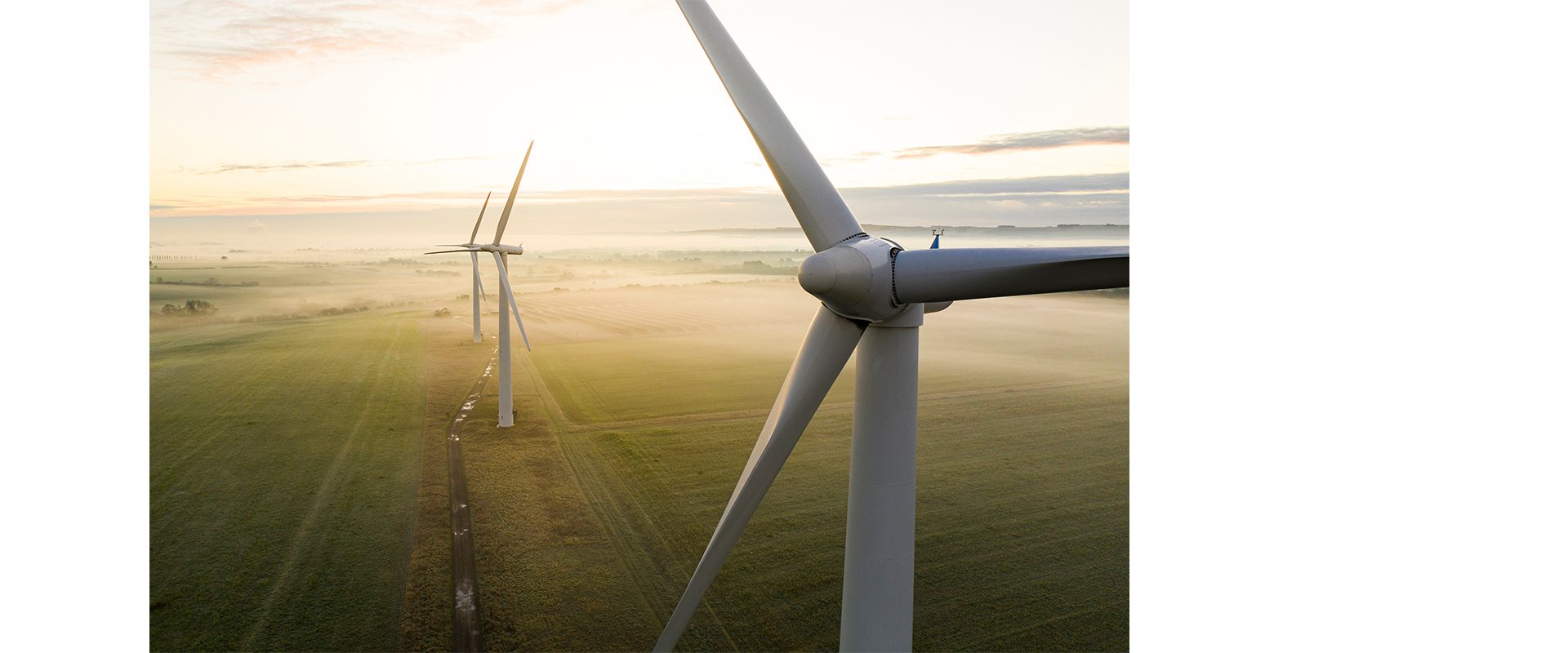 Two wind turbines standing in a green field during early morning, with a soft mist covering the ground and a light gradient in the sky.